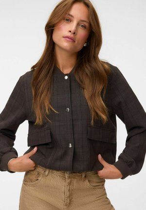 Young woman with long brown hair wearing a dark buttoned jacket and beige pants, standing with hands in jacket pockets against white background.