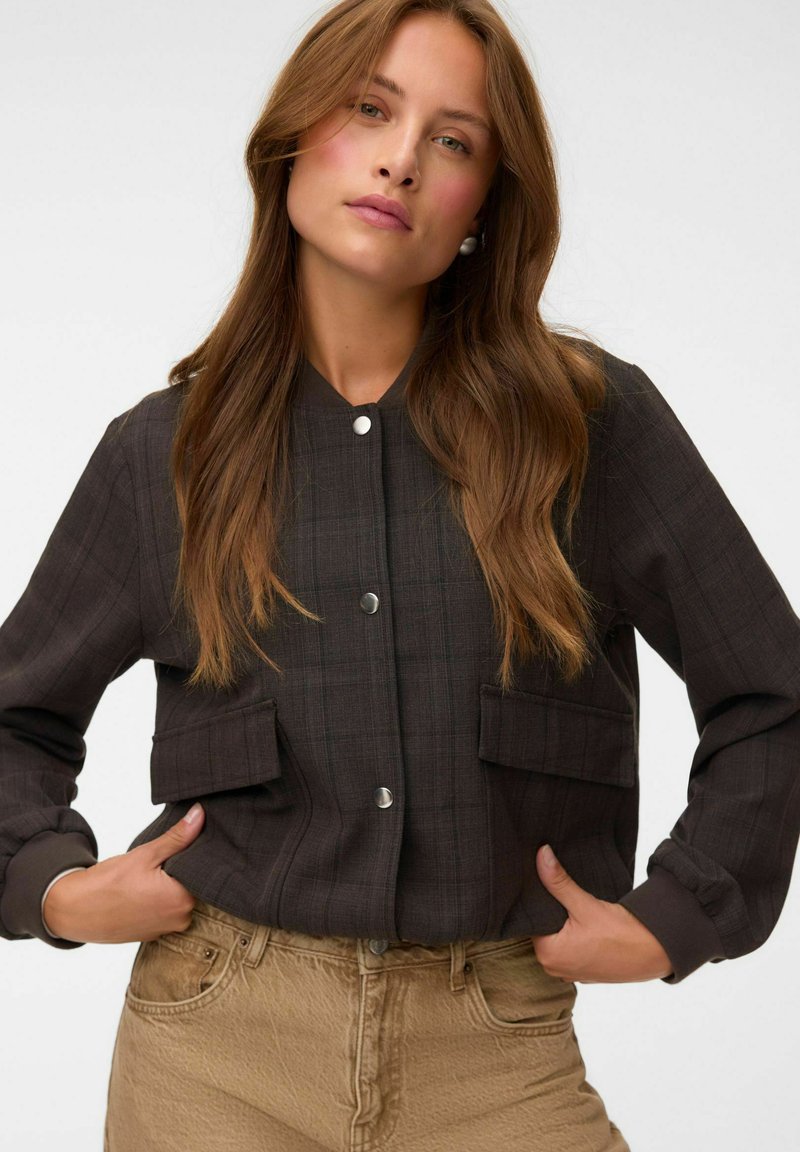 Jeune femme aux longs cheveux bruns, portant une veste foncée boutonnée et un pantalon beige, debout avec les mains dans les poches de la veste sur fond blanc.