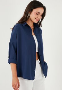 Camicia button-up blu navy realizzata in tessuto morbido, con colletto, maniche arrotolate e una vestibilità rilassata, infilata in pantaloni di colore chiaro.