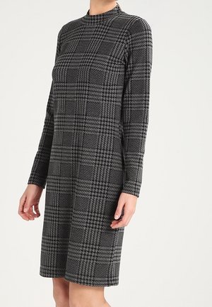 Femme portant une robe longueur genou, manches longues, col montant, avec un motif pied-de-poule noir et gris sur un fond clair.