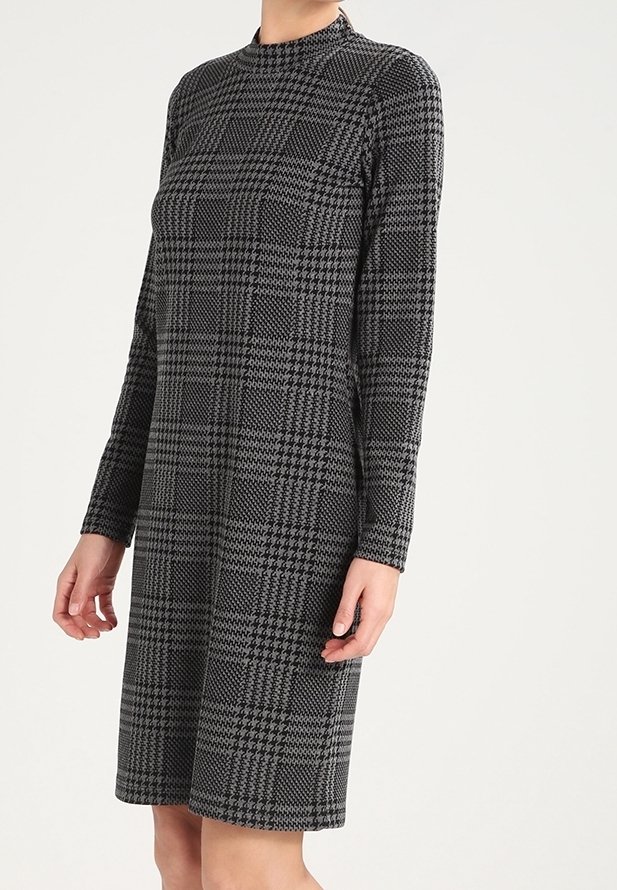 Femme portant une robe longueur genou, manches longues, col montant, avec un motif pied-de-poule noir et gris sur un fond clair.