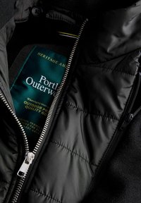 Czarna kurtka puchowa o błyszczącym wykończeniu, pikowanej teksturze, srebrnym zamku błyskawicznym i zielonej metce z napisem "Portland Outerwear" wewnątrz.