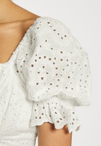 Blusa branca com tecido texturizado, bordados em ilhós, mangas bufantes e bainha ondulada com pregas.