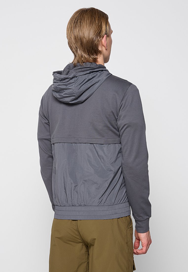 Uomo che indossa una giacca con cappuccio di un grigio scuro con pannello posteriore in nylon e pantaloni verde oliva, visto da dietro su uno sfondo semplice.