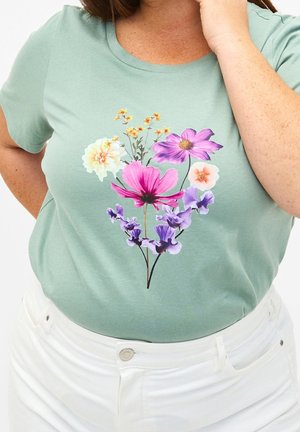 Kvinde iført salviegrøn t-shirt med farverigt blomstermønster og hvide bukser, hånd hvilende nær hendes ansigt.