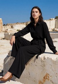 Mujer de cabello oscuro que lleva un mono negro de manga larga y tacones marrones, sentada sobre un gran bloque de piedra en una zona rocosa iluminada por el sol.