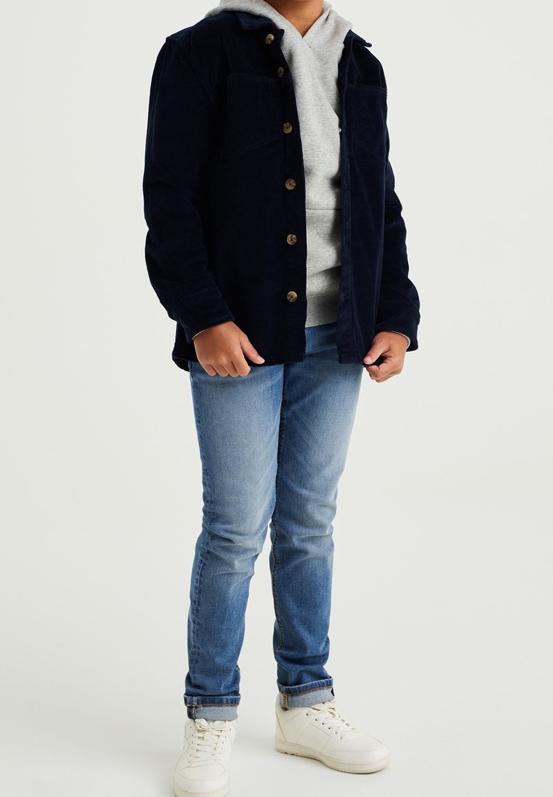 Námořnická kordová košile-jacket s dvěma náprsními kapsami, nošená přes šedou mikinu, spárovaná se světle modrými džíny a bílými teniskami.