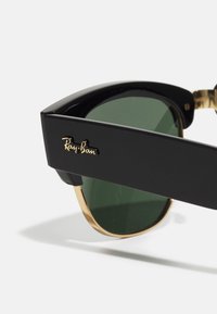 Ochelari de soare Ray-Ban cu rame din plastic negru, lentile verzi și accente aurii pe brațele ochelarilor. Prezintă un logo scris cursiv pe lateral.
