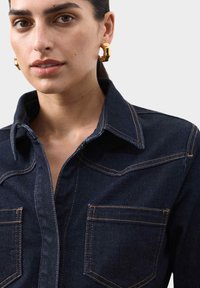 Mørk denimskjorte med spids krave, to brystlommer og kontrastsyning. Bemærkelsesværdige guldringe bæres som accent.