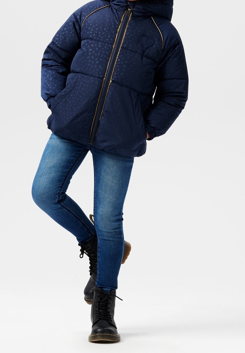 Enfant portant une veste matelassée bleu marine, un jean skinny bleu et des bottes noires à lacets, debout avec une jambe croisée sur l'autre.