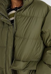 Chaqueta acolchada de color verde oliva con un alto cuello, cierre a presión y costuras visibles. Presenta un bolsillo delantero y una textura de tela suave y brillante.