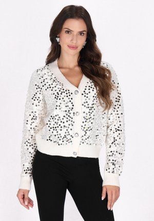 Weißer gestrickter Cardigan, verziert mit silbernen und schwarzen Pailletten, mit V-Ausschnitt, Knopfverschluss und gerippten Bündchen. Unter der Jacke werden schwarze Hosen getragen.