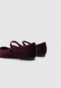 Bailarinas de terciopelo burdeos con punta redondeada y cierre de doble tira. Los zapatos tienen una suela baja de goma negra y una textura suave.