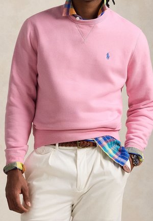 Mann trägt rosa Sweatshirt über buntem kariertem Hemd mit weißer Hose und braunem geflochtenem Gürtel, Hand in der Tasche, Uhr am linken Handgelenk.