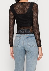 Top in pizzo nero a maniche lunghe con un design aderente e scollatura rotonda, sovrapposto a una maglietta nera a tinta unita, abbinato a jeans azzurri chiari.