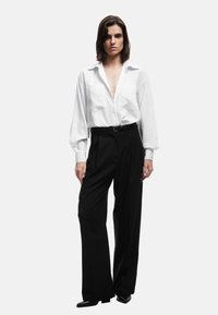 Camiseta blanca a rayas con un escote profundo y dos bolsillos en el pecho, combinada con pantalones negros de cintura alta y zapatos negros elegantes.