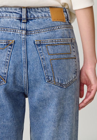 Vaqueros de mezclilla azul claro con dos bolsillos traseros con costuras amarillas y una etiqueta de cuero marrón con la palabra "CLAUDIE" en la cintura.
