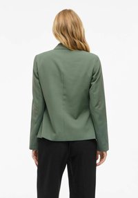 Blazer verde de manga larga, diseño entallado, textura suave y solapa con muesca, combinado con pantalones negros.