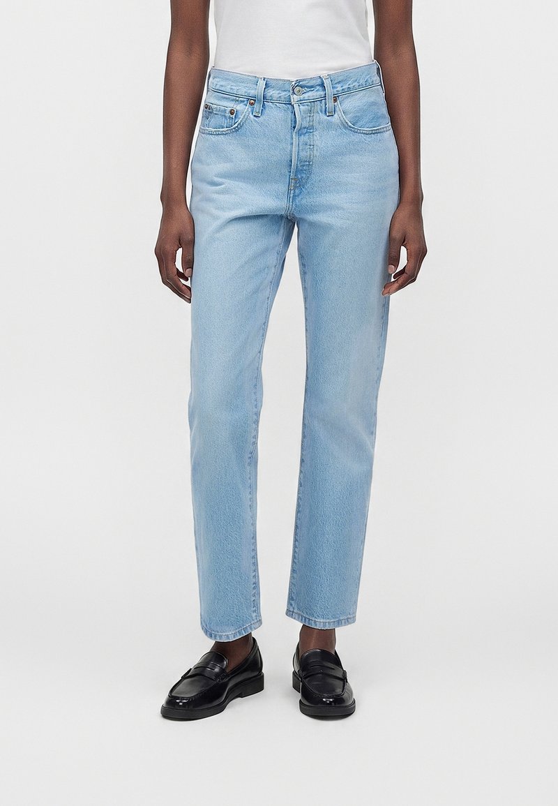 Personne portant un jean bleu clair droit, un haut blanc et des mocassins en cuir noirs, se tenant devant un fond uni.