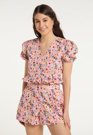 Jonge vrouw lacht terwijl ze een roze korte bloemenoverall draagt met opgepofte mouwen en ruches in de taille.