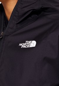 Zwarte jas met een ritssluiting aan de voorkant en een verstelbare capuchon. Heeft een opvallend wit logo aan de linkerkant. Gestructureerde stof met een soepele afwerking.