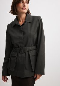 Dark gray belted jacket with a collar, featuring a structured fit, large front pockets, and subtle texture. Material appears smooth and durable.