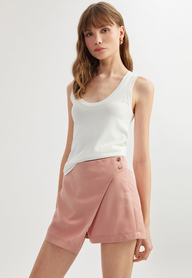 Débardeur blanc sans manches associé à un short rose clair enroulé avec des accents de boutons et une texture douce et lisse.
