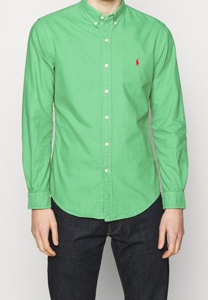 Mężczyzna ubrany w jaskrawozieloną koszulę na guziki z długim rękawem z czerwonym haftowanym logo, w ciemnoniebieskich dżinsach, stojący na jednolitym tle.