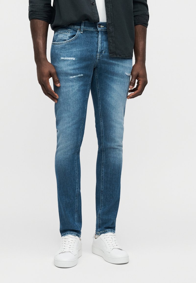 Jean en denim bleu avec des décolorations légères et des détails usés sur les cuisses, assorti à des baskets blanches. Coupe slim et taille moyenne.