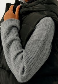 Černá péřová vesta se stříbrným kováním, vrstvená přes světle šedý ribbovaný vlněný svetr. Texturovaná tkanina kontrastuje s lesklým materiálem vesty.