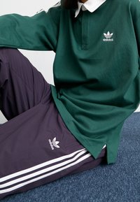 Grön långärmad pikétröja med vit krage och Adidas-logotyp; kombinerad med mörklila byxor med vita sidostripes och logotyp.