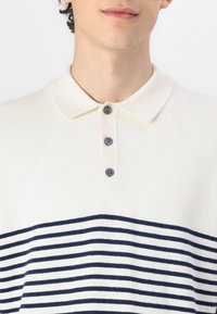 Polo à manches courtes crème avec des rayures horizontales bleues marine, fabriqué en tissu doux. Il présente un col et trois boutons foncés à l'avant.