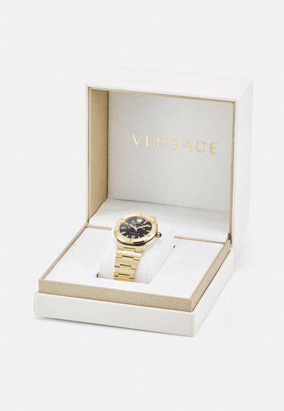 Gold-colored Versace watch in a white box with soft white padding. Features a black dial and gold-toned linked bracelet. Gold accents visible.