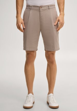 Mężczyzna ubrany w beżowe krótkie spodenki do kolan z przednimi kieszeniami i białe trampki, stojący na tle jednolitego jasnego tła.