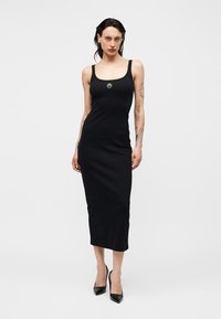 Kobieta ubrana w czarną sukienkę midi bez rękawów z okrągłym dekoltem i czarne szpilki, stojąca na tle jednolitego jasnego tła.