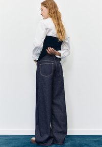 Ženska z dolgimi blond lasmi, oblečena v belo srajco in črn brezrokavnik, stoji bok ob boku v širokih temnih denim hlačah na modrem preprogi.