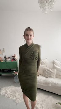 Femme aux cheveux clairs portant une robe moulante à manches longues vert olive, debout dans une pièce avec un canapé blanc, un meuble vert et un tapis moelleux.