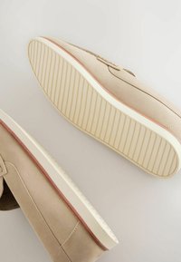 Beige Wildleder-Sneaker mit einer strukturierten Gummisohle, ausgestattet mit einer hellcremefarbenen Zwischensohle und einem dünnen braunen Akzent am Rand.
