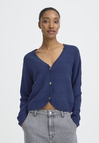 Mørkeblå cardigan med V-hals, ribbet struktur og bølget kant. Har fem metalliske knapper og et let cropped design.