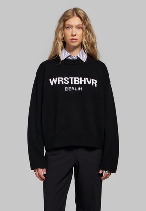 Femme blonde debout devant un fond gris, portant un pull noir avec le texte "WRSTBHVR BERLIN", une chemise rayée à col et un pantalon noir.