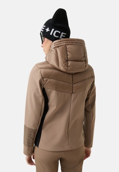 Osoba w ocieplanej brązowej kurtce z kapturem i czarnej beanie z białym napisem, widziana z tyłu na jednolitym tle.