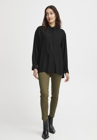 Camicia nera a maniche lunghe con bottoni e colletto, abbinata a pantaloni verdi slim-fit e stivali neri alla caviglia. Tessuto liscio, vestibilità casual.
