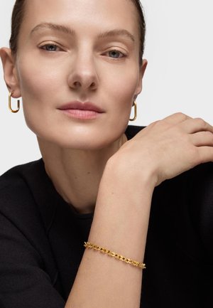 Kobieta z krótkimi włosami, nosząca złote kolczyki-kółka i złotą bransoletkę na łańcuszku, opierająca ramię przy twarzy na jednolitym tle.