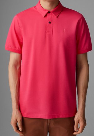 Man draagt een felroze korte mouw polo met twee zwarte knopen en een bruine broek, tegen een grijze achtergrond.