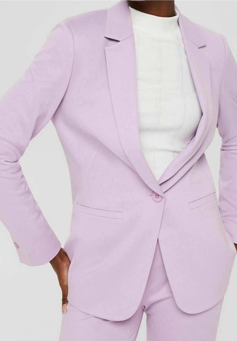 Blazer violet clair en tissu lisse, avec un col cranté, une fermeture par un seul bouton et des poches latérales, associé à un haut blanc.