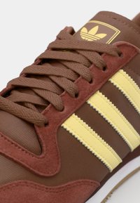 Brune sneakers med overdel av semsket skinn og tekstil, med tre gule striper. Tykke brune lisser og en logo med gulldetaljer på tungen.
