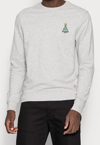 Felpa grigio chiaro con un albero di Natale verde ricamato e una stella gialla. Polsini e orlo a costine, texture liscia.