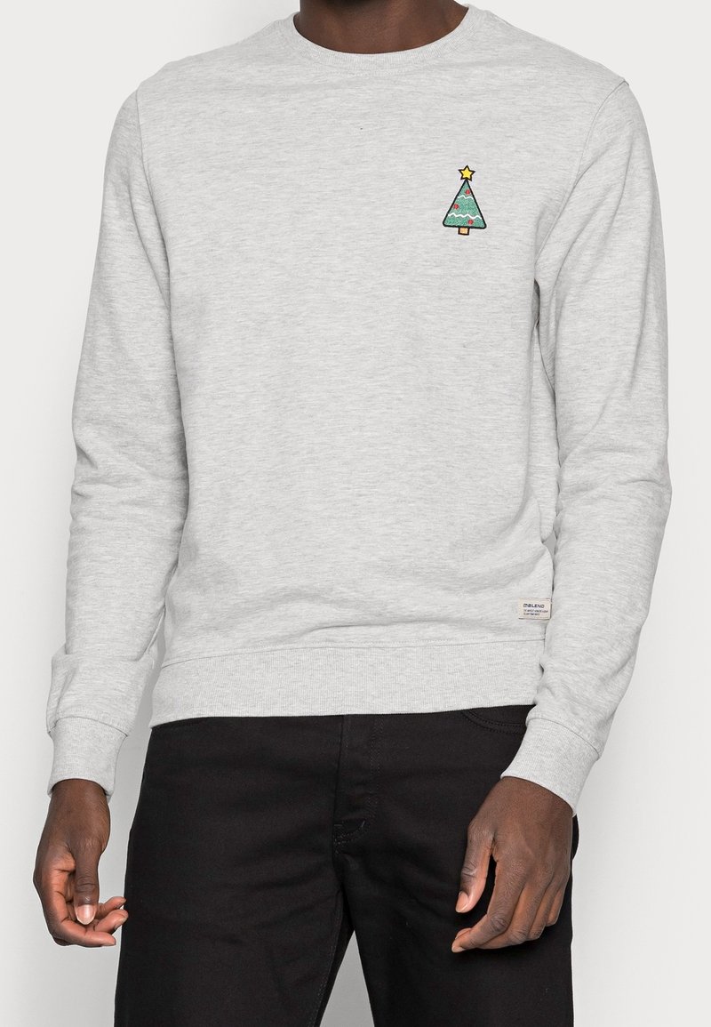 Felpa grigio chiaro con un albero di Natale verde ricamato e una stella gialla. Polsini e orlo a costine, texture liscia.