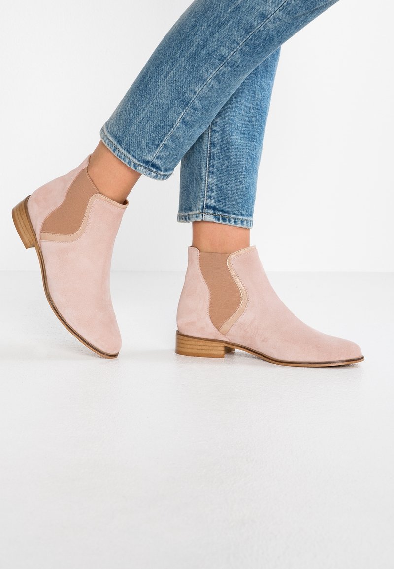 Personne portant des bottines en daim rose clair avec des panneaux élastiques sur les côtés et des talons bas en bois, associées à un jean en denim bleu sur fond blanc.