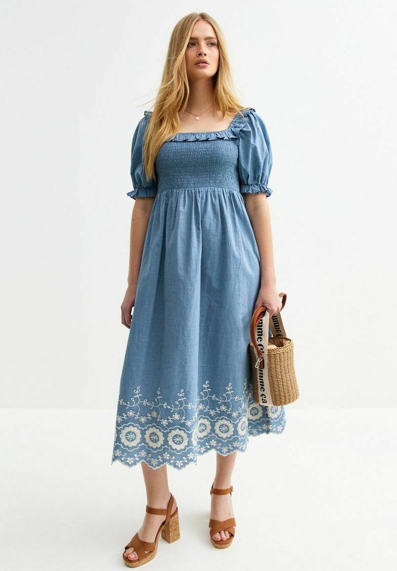 Vestido midi de mezclilla azul con mangas abullonadas, cuerpo fruncido, bordado floral en el dobladillo y borde con forma de ondas. La modelo sostiene un bolso de mimbre.